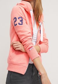 Donna che indossa una felpa rosa con zip, con il numero 23 sulla manica e un'etichetta del logo Superdry, abbinata a jeans scuri e una maglietta bianca.
