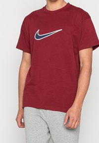 Hnědá bavlněná tričko s kulatým výstřihem. Na hrudi se nachází velké bílé a námořnické logo Nike swoosh. Krátké rukávy a uvolněný střih.