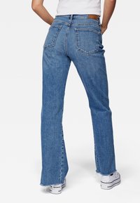 Persona que lleva jeans rectos de cintura alta en azul con dobladillo deshilachado y zapatillas blancas, de pie con las manos en las caderas.