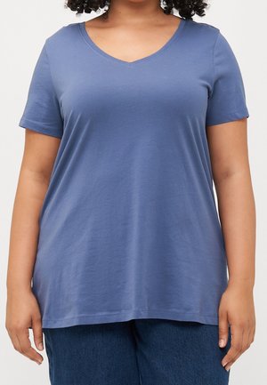 Person, die ein kurzes blaues V-Ausschnitt-T-Shirt und dunkelblaue Hose trägt, steht vor einem schlichten hellen Hintergrund.