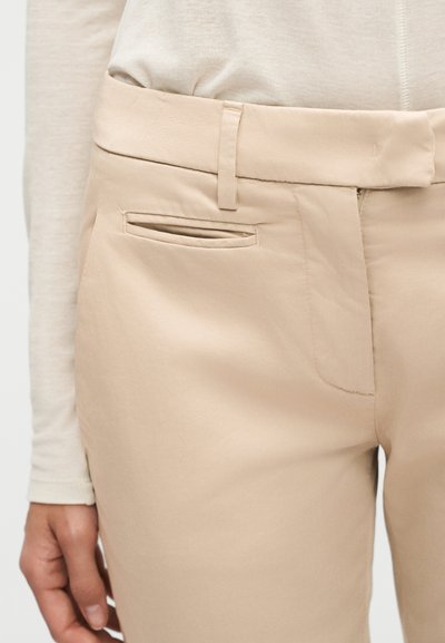 Μπεζ παντελόνι σε στενή γραμμή με ζώνη στη μέση, θηλιές για ζώνη, τσέπη με σχισμή και ένα χέρι που ακουμπά δίπλα του, σε συνδυασμό με μια κρεμ μακρυμάνικη μπλούζα.