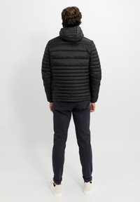 Giacca imbottita nera con cappuccio, caratterizzata da un design trapuntato orizzontale e polsini elasticizzati. Indossata sopra pantaloni blu navy e sneakers bianche.