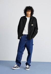 Black button-up shirt, white t-shirt underneath, blue denim jeans, and black sneakers with white accents. Model stands against a plain background.