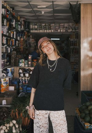 Osoba ubrana w czarną, oversize'ową koszulę i białe, koronkowe spodnie stoi przed sklepem pełnym różnych produktów, w tym warzyw.