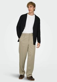 Mustast kootud kardigan nööpidega valge särgi peal, kombineeritud beežide volditud pükste ja pruunide jalanõudega. Sile tekstuur ja lõdvestunud lõige.