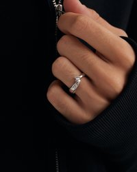 Anillo de plata con un diseño suave y retorcido, exhibido en una mano que sostiene una prenda negra con cremallera. Tela negra texturizada visible.