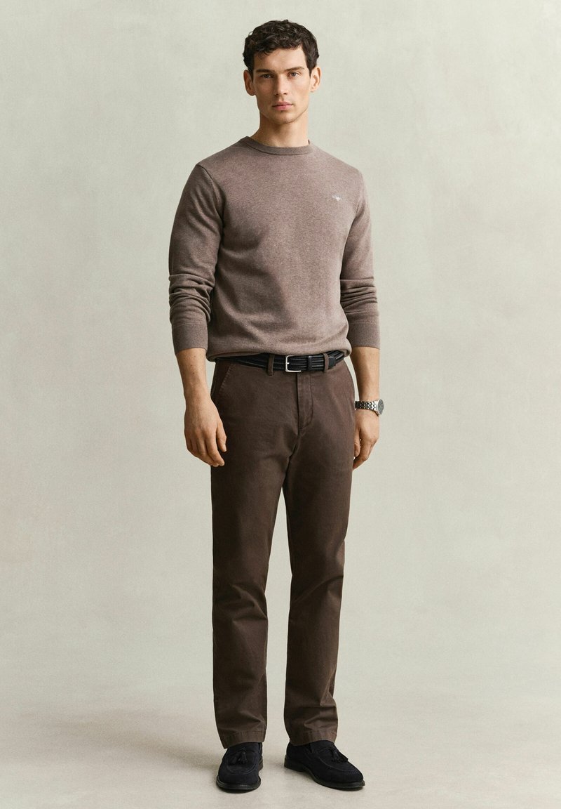 Junger Mann, der einen braunen Pullover mit langen Ärmeln, dunkelbraune Hose, schwarzen Gürtel, schwarze Schuhe und eine Armbanduhr vor einem einfarbigen Hintergrund trägt.