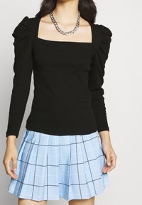 Svart långärmad topp med fyrkantig urringning och puffiga axlar, i kombination med en veckad ljusblå kjol med svarta rutmönster.