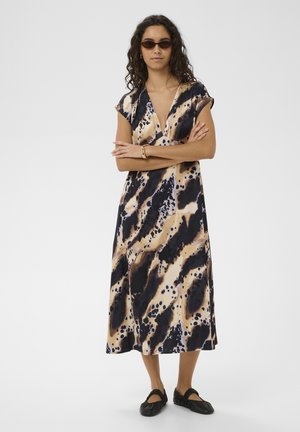 Frau mit lockigem Haar, die ein braun-schwarz gemustertes Midikleid, dunkle Sonnenbrillen, schwarze Ballerinas und ein goldenes Armband trägt, mit verschränkten Armen steht.