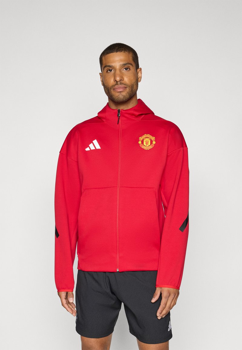 Crvena jakna na zip s kapuljačom, s logom Manchester Uniteda, bijelim Adidas trakama i crnim detaljima na rukavima. Crne kratke hlače.