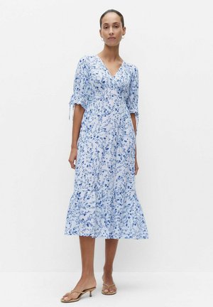 Kobieta w jasnoniebieskiej sukience w kwiaty do kolan z półrękawami i beżowymi sandałami na obcasie, stoi na białym tle.