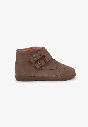 Stivale alla caviglia in suede marrone con doppia fibbia, punta rotonda, piccoli dettagli perforati e fodera interna in contrasto color sabbia. Suola in gomma piatta.