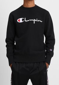Felpa nera con colletto rotondo, con logo "Champion" ricamato in bianco. Texture liscia e polsini e orlo a coste.