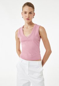 Top sin mangas de cuello en V de color rosa con un diseño ajustado y textura acanalada, acompañado de pantalones blancos de cintura alta, sobre un fondo liso.