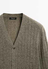 Stickad cardigan i jordnära bruna nyanser, med V-ringning, ribbad textur och två knappar framtill. Mjuk tyg med ett subtilt mönster.