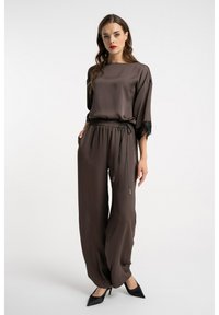 Brązowy satynowy kombinezon z szerokimi nogawkami i dopasowaną talią. Ozdobiony koronkowymi akcentami na rękawach i ze ściągaczem w pasie. Do kompletu czarne buty.