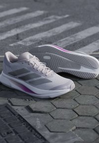 Hellgraue Sportschuhe mit schwarzen Streifen und violetten Akzenten, die auf sechseckigem Pflaster nahe einem Fußgängerüberweg liegen.