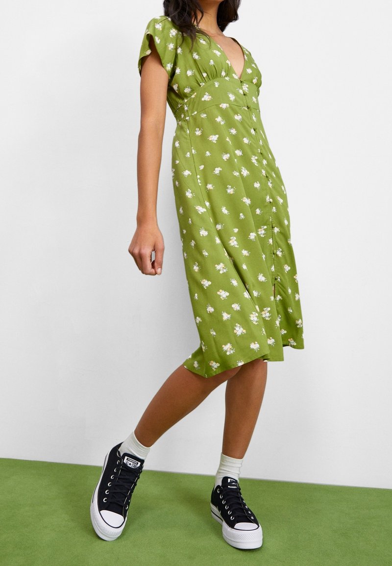 Femme portant une robe midi verte à fleurs avec des manches courtes, assortie de chaussettes blanches et de baskets compensées noires, debout sur un sol vert.