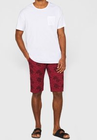 Homme portant un t-shirt blanc à manches courtes, un short rouge avec un motif de palmiers, et des sandales noires, se tenant avec une main dans la poche.