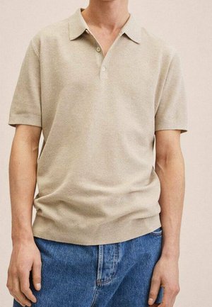 Persoon draagt een beige polo met korte mouwen en blauwe jeans, staat met ontspannen armen langs het lichaam tegen een neutrale achtergrond.