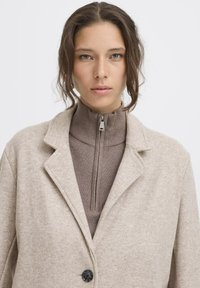 Beige wollen blend jas met notch-lapels, enkele zwarte knoop, gedragen over een bruine hoge hals met rits. Smeuïge textuur, getailleerde pasvorm.