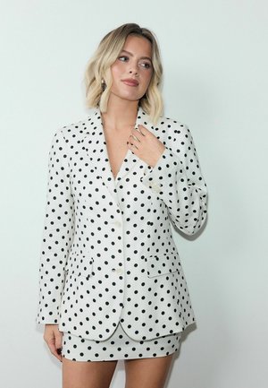 Blondynka w białej marynarce i spódnicy w czarne groszki, pozująca na jednolitym jasnym tle, z ręką przy kołnierzu.