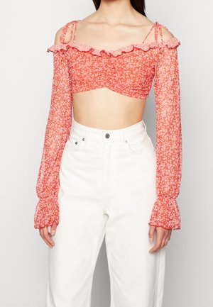 Kobieta ubrana w czerwony kwiatowy, odkrywający ramiona krótki top z długimi przezroczystymi rękawami oraz wysokim stanem białe spodnie na jednolitym białym tle.