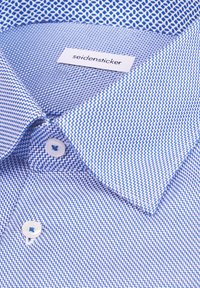 Chemise à motifs bleus avec une texture en tissage, boutons blancs, et une étiquette "seidensticker" sur le col.