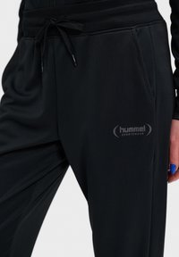 Zwarte sportbroek met een zachte textuur, voorzien van een trekkoord in de taille, zijzakken en een grijs Hummel-logo op de linkerdij.