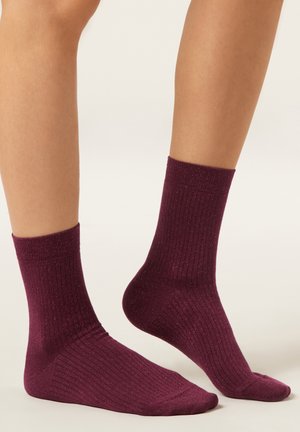 Bordeauxrote, gerippte Socken mit weicher Textur und dezentem Glanz. Die kurze Länge reicht knapp über den Knöchel und sorgt für eine eng anliegende Passform.