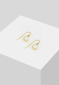 Boucles d'oreilles à crochet en or avec des pierres blanches rondes, présentées sur une surface blanche inclinée contre un fond gris clair.