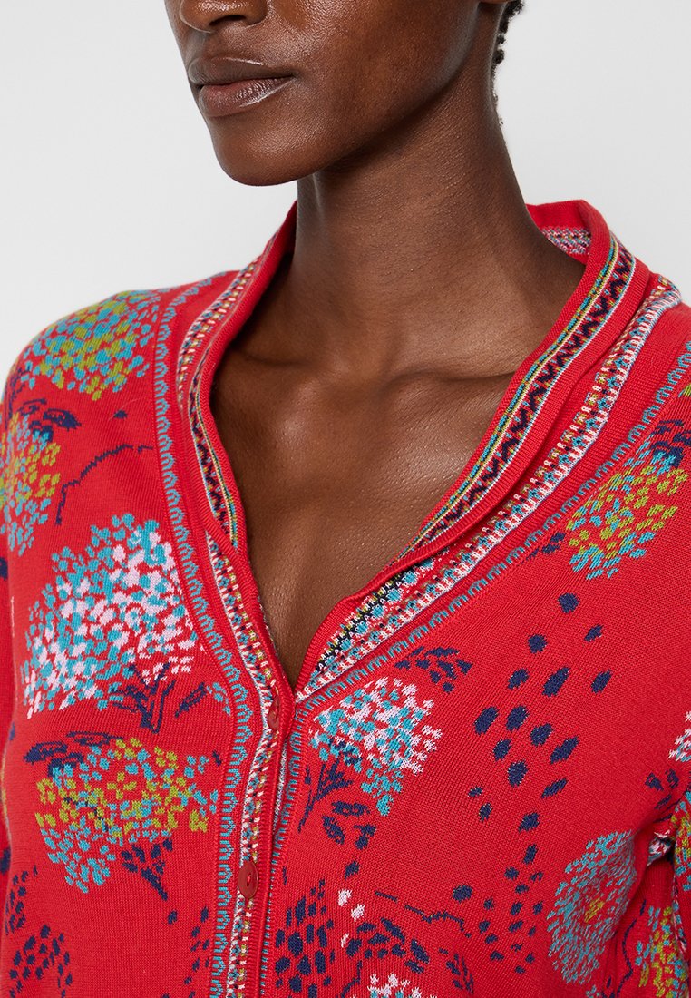 Czerwony kardigan z kwiecistym wzorem w odcieniach niebieskiego, zielonego i białego. Posiada dekolt w serek, zapięcie na guziki oraz misternie wykończone detale.