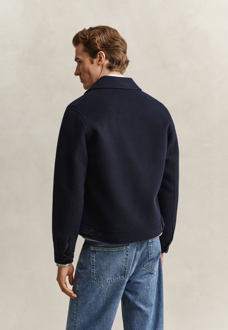 Uomo con capelli castano chiaro che indossa una giacca blu scuro e jeans blu, in piedi di profilo su uno sfondo semplice.