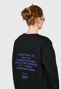 Kobieta z brązowymi włosami związanymi w kok, nosząca białe okulary przeciwsłoneczne i czarną bluzę z niebieskim tekstem o tematyce taktycznej na plecach.