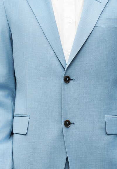 Ανοιχτό μπλε σακάκι με δύο μαύρα κουμπιά κουμπωμένα πάνω από ένα λευκό πουκάμισο με κουμπιά, που δείχνει λεπτομέρειες από το μπροστινό μέρος του κορμού.