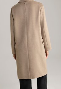 Béžový vlněný kabát s dlouhými rukávy, rovného střihu a zadním rozparkem. Má jemnou texturovanou úpravu a knoflíkový detail u krku.