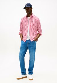 Camicia a righe rossa e bianca con bottoni, t-shirt bianca, jeans blu e sneakers bianche con suola marrone, completati da un cappellino da baseball blu navy.