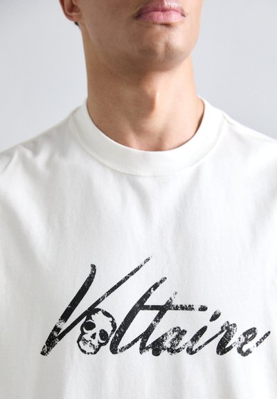 Camiseta de algodón blanca con un gráfico negro que presenta el texto "Voltaire" y un diseño de calavera. Cuello redondo clásico con un ajuste relajado.