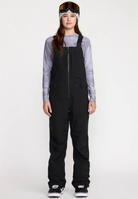 Salopette de ski noire isolante avec bretelles réglables, fermeture éclair avant et multiples poches, associée à un haut à manches longues violet clair.