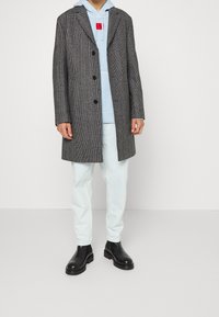 Abrigo gris de espiga con solapas de muesca, botones negros, superpuesto a una sudadera de color azul claro, combinado con jeans de denim claros y botas negras.