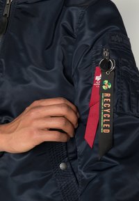 Giacca bomber blu navy con una trama liscia. Presenta una tasca con un portachiavi nero e tag in tessuto rossi e neri con la scritta "RICICLATO."