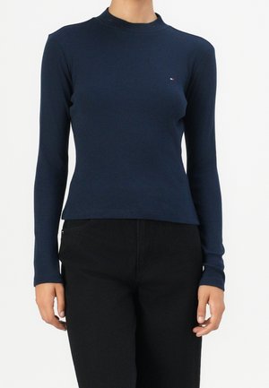 Kobieta ubrana w dopasowaną, granatową bluzkę z długim rękawem z małym logo na piersi i czarne spodnie na jednolitym tle.