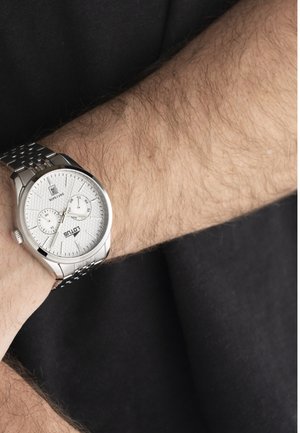 Moška srebrna kovinska zapestna ura z belim številčnico, podštevilčnicama za dan in datum, nošena na poraščeni roki preko črnega rokava.