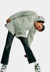 Giacca leggera verde chiaro con cappuccio, chiusura a bottoni e tasche frontali, abbinata a jeans scuri e sneakers con dettagli verdi.