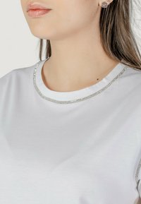 Camiseta blanca con escote redondo y un ribete de strass plateado. Material suave, forma ajustada, sin patrones visibles ni adornos adicionales.