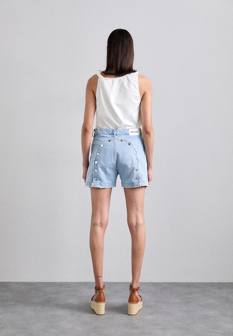 Shorts di jeans blu chiaro con dettagli in bottoni lungo i lati, orlo grezzo e una canotta bianca. Gli espadrilles marroni con zeppa completano l'outfit.