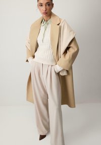 Beige villakangas takki, jossa on jämäkkä siluetti, kermaista palkinengedonia neuletta, vaaleanvihreä kaulussolki ja leveät vaaleat housut, joissa on kiiltävä pintakuvio.