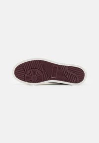 Rubber sole with a maroon bottom, white edging, and textured grip pattern. Features a circular accent and embossed logo detail.