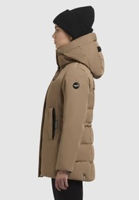 Khaki värvi puhuri jakk kõrge kraega, lukuga ees ja küljetaskutega. Must logopatch varrukal ja tekstuuriga must müts.