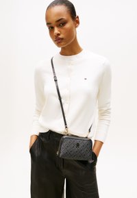 Femeie îmbrăcată într-un cardigan alb cu nasturi și pantaloni din piele negri, cu mâinile în buzunare, purtând o geantă mică de umăr cu un model închis.